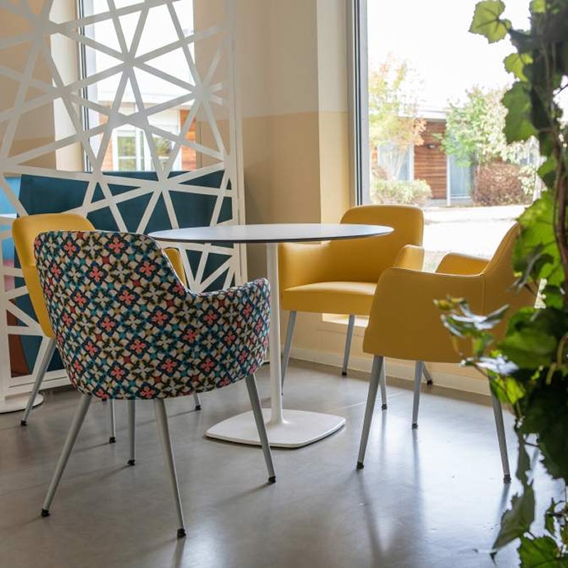 Coin salon moderne : chaises jaunes et table de pause Coin salon moderne avec table ronde et quatre chaises : une à motif multicolore et trois jaunes unies, près d'une fenêtre avec paravent géométrique.