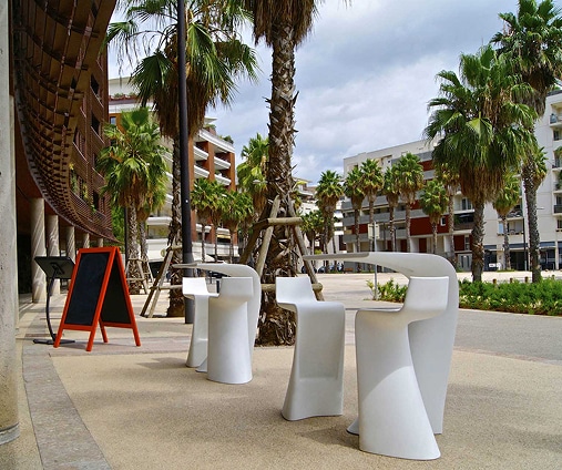 Mobilier de terrasse design sous palmiers, ambiance méditerranéenne Mobilier de terrasse blanc design sous palmiers, avec un tableau noir sur chevalet orange près d'un bâtiment moderne.