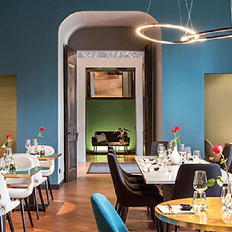 Restaurant Chic Design : Atmosphère Élégante et Moderne Restaurant chic aux murs bleu sarcelle, tables dressées, chaises modernes blanches et noires, éclairage design circulaire au plafond.