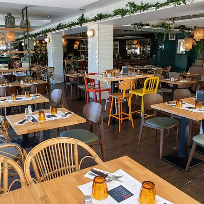 Restaurant branché: chaises colorées et ambiance végétale Restaurant moderne avec tables en bois clair, chaises colorées (jaune, rouge) et rotin, décoré de plantes vertes en suspension. Menu "PASTA" visible.