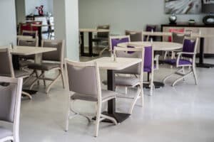 Salle à manger moderne et lumineuse avec tables carrées et rondes, chaises taupe et violettes, et sol clair.